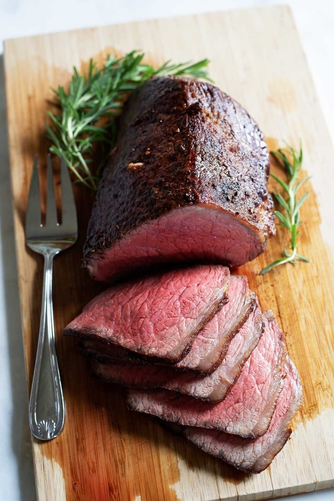 roast beef sliced on cutting board with rosemary and serving fork, how to cook melt in the mouth roast beef