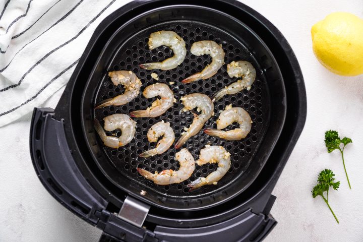 raw shrimp in black air fryer basket with white background and dish towel